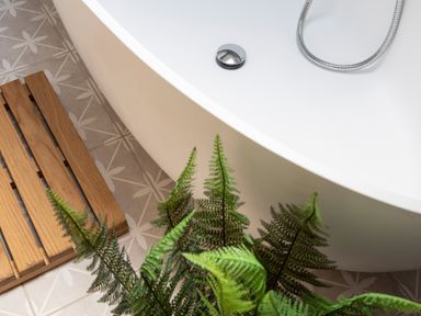 A bathroom corner with a white bathtub a wooden bath mat patterned tiles and green fern leaves at Gatesgarth in Coniston