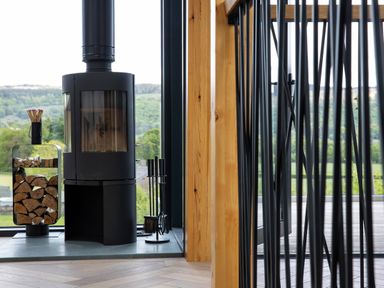A black wood-burning stove with glass windows beside a wood holder and fireplace tools near large windows showing a green landscape at Gatesgarth in Coniston
