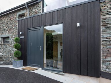 A modern entrance with a gray door and large window next to a stone wall with a potted topiary at Gatesgarth in Coniston