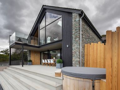 A modern house exterior with large glass windows wooden and stone walls outdoor dining table and covered hot tub at Gatesgarth in Coniston