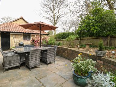 An outdoor patio with a glass table and wicker chairs under an umbrella next to a garden and a small building at Beckhythe Cottage in Overstrand