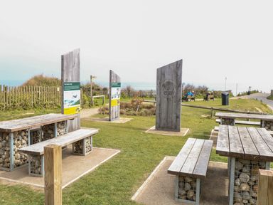 An outdoor area with wooden picnic tables and benches with stone bases and wooden information boards on grass near a pathway and fence at Beckhythe Cottage in Overstrand