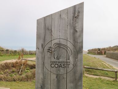 A wooden sign with the words Deep History Coast and mammoth logo near a road and grassy area at Beckhythe Cottage in Overstrand