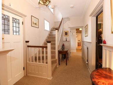 A hallway with stairs a small wooden table framed pictures and a door with decorative glass panels at Beckhythe Cottage in Overstrand