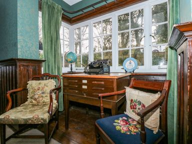 A corner room with two wooden chairs with floral cushions a wooden desk with drawers a typewriter a globe and a decorative plate by windows with green curtains at Beckhythe Cottage in Overstrand
