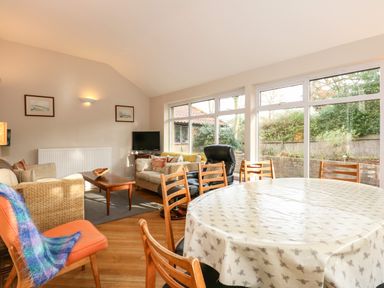 A living room with a round dining table and wooden chairs near large windows at Beckhythe Cottage in Overstrand