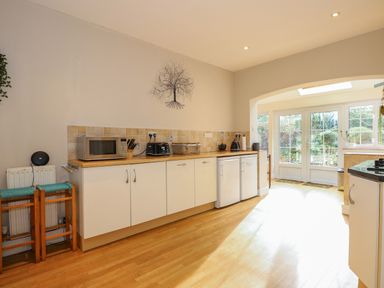 A kitchen with white cabinets a microwave a toaster and two small fridges leading to a sunlit area with glass doors at Beckhythe Cottage in Overstrand