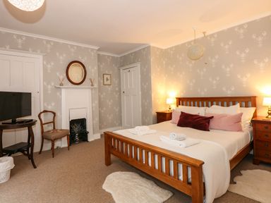 A bedroom with a wooden bed with white linens pillows bedside tables lamps a chair a TV a white fireplace and grey lobster wallpaper at Beckhythe Cottage in Overstrand