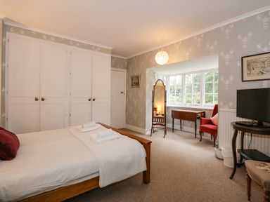 A bedroom with a wooden bed, white linens, built-in white wardrobes, a standing mirror, a wooden table, a red armchair, and a small TV on a round table at Beckhythe Cottage in Overstrand