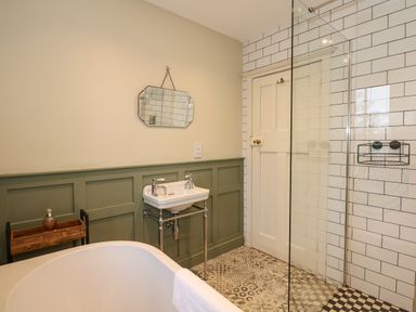 A bathroom with a bathtub a sink a mirror a wooden shelf a door and a tiled shower area at Beckhythe Cottage in Overstrand