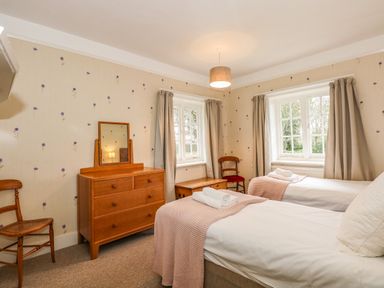 A bedroom with two single beds, wooden dresser with mirror, two chairs, and two windows with curtains at Beckhythe Cottage in Overstrand
