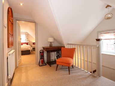 A hallway landing with an orange chair a wooden desk with a lamp a fire extinguisher and a view into a bedroom at Beckhythe Cottage in Overstrand