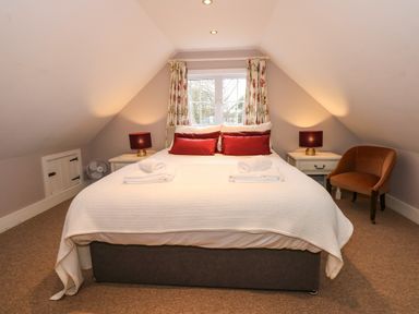 A bedroom with a double bed red pillows two bedside tables with lamps a brown chair and floral curtains at Beckhythe Cottage in Overstrand