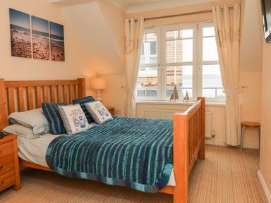 A bedroom with a wooden bed covered with a blue striped blanket and pillows next to a window with cream curtains at Beach Lodge in Woolacombe