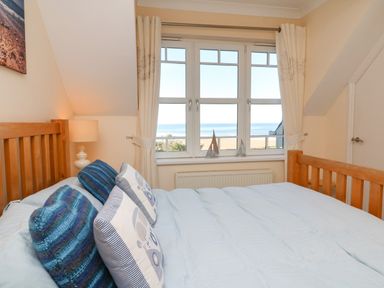 A bedroom with a wooden bed and pillows facing a window with curtains and a beach view at Beach Lodge in Woolacombe