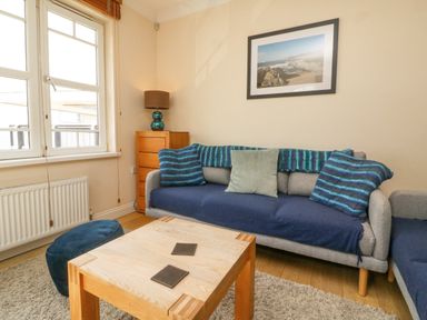 A living room with a grey sofa with blue and green cushions a wooden coffee table a chest of drawers with a lamp and a framed picture on the wall at Beach Lodge in Woolacombe