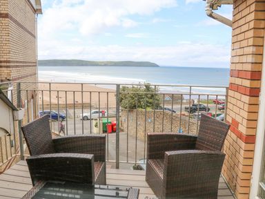 A balcony with two wicker chairs and a glass table overlooking a beach and ocean at Beach Lodge in Woolacombe