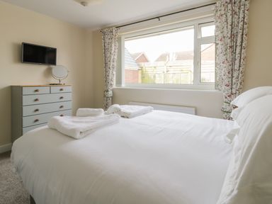 A bedroom with a bed covered in white linens and towels on top a dresser with a round mirror and a wall-mounted TV a window with floral curtains at Arbour Retreat in Filey
