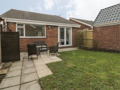 A backyard with a paved patio area, outdoor chairs, a small table with a bottle and glasses, grass lawn, and a brick house with white framed windows and a door at Arbour Retreat in Filey
