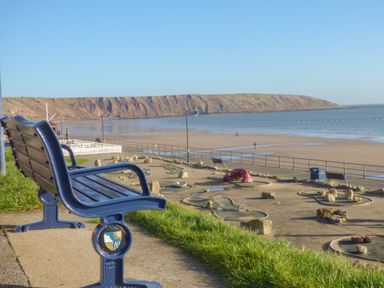 A blue bench overlooking a mini golf course with the beach and cliffs in the background at Arbour Retreat in Filey