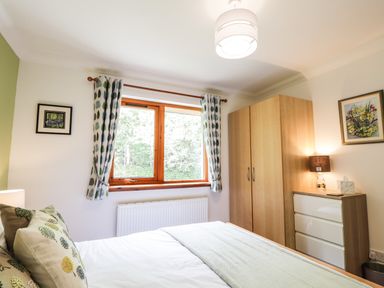 A bedroom with a bed pillows curtains a window a wardrobe a chest of drawers a lamp and framed pictures at Silver Birch Lodge in Ratagan Glenshiel near Dornie