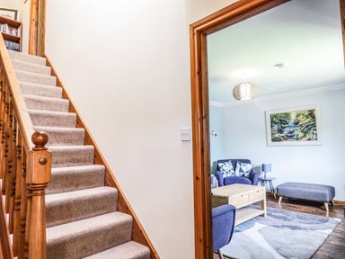 A carpeted staircase with wooden railing next to a living room with blue chairs a wooden coffee table and a gray ottoman at Silver Birch Lodge in Ratagan Glenshiel near Dornie