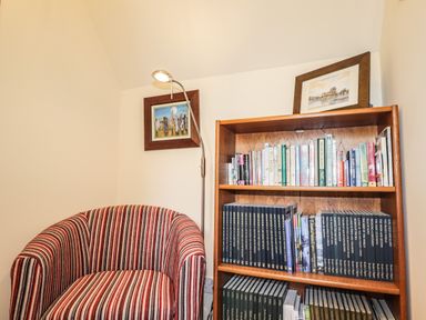 A corner with a striped armchair a floor lamp and a wooden bookshelf filled with books and framed paintings on the wall and shelf at Silver Birch Lodge in Ratagan Glenshiel near Dornie