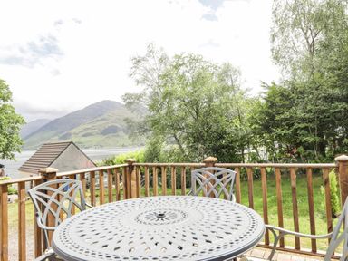 A patio with metal table and chairs overlooking trees a roof and mountains at Silver Birch Lodge in Ratagan Glenshiel near Dornie