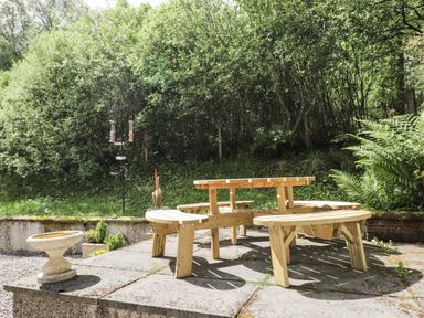 A wooden picnic table with attached benches on a stone patio beside a bird feeder and greenery at Silver Birch Lodge in Ratagan Glenshiel near Dornie