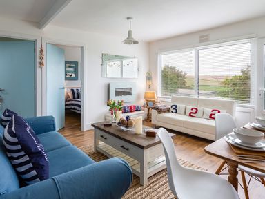 A living room with blue and white sofas a wooden coffee table white chairs and a dining table at The Hut in Dartmouth