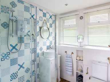 A bathroom with a shower enclosure featuring blue patterned tiles a round mirror a white sink two windows with blinds and a toilet at The Hut in Dartmouth