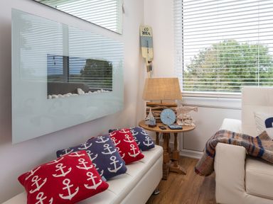 A living room with white sofas red and blue anchor patterned cushions a wooden side table with a lamp and decorative items and a large wall mounted electric fireplace at The Hut in Dartmouth