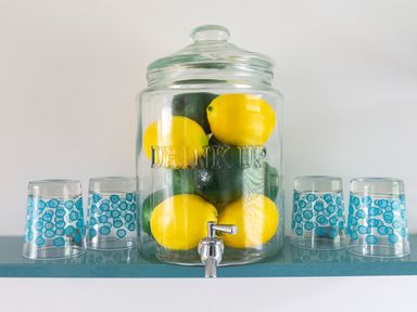 A glass dispenser filled with lemons and limes with four clear glasses with blue circular patterns on a shelf at The Hut in Dartmouth