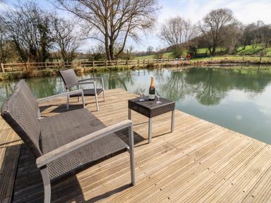 A wooden deck with two chairs a small table with a bottle and two glasses by a pond at Acorn Lodge At Bridge Lake Farm & Fishery in Chacombe