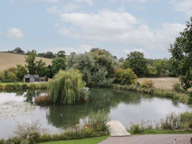 A pond with a small dock and willow trees surrounded by fields and bushes at Acorn Lodge At Bridge Lake Farm & Fishery in Chacombe