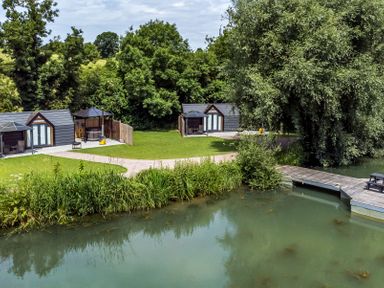 A lakeside view with two small cabins a grassy area and trees at Acorn Lodge At Bridge Lake Farm & Fishery in Chacombe