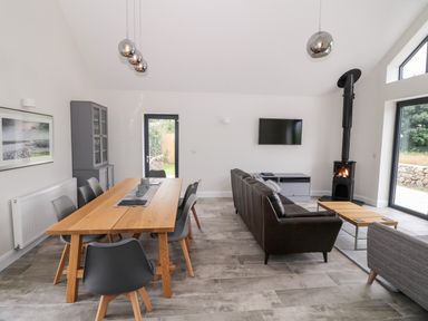 A living room and dining area with a wooden dining table and gray chairs a brown sofa a TV mounted on the wall and a wood stove at Pen Y Cefn in Pen Y Cefn