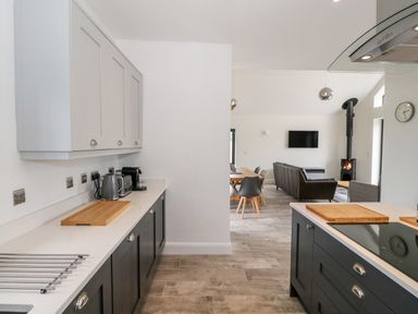 A kitchen with cabinets appliances and cutting boards opening to a living room with dining table chairs sofa wall mounted tv and wood stove at Pen Y Cefn
