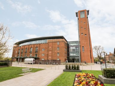 A large brick building with a tall tower and a glass entrance with rsc welcome sign near a path with flowers and benches