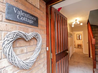 An open wooden front door with a heart-shaped wreath and a sign reading Valentines Cottage leading to a hallway with carpet and stairs at Valentines Cottage Stratford-Upon-Avon