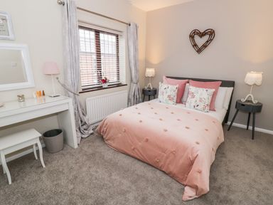 A bedroom with a bed pink bedding floral pillows side tables lamps window with curtains and a dressing table at Valentines Cottage in Stratford-Upon-Avon