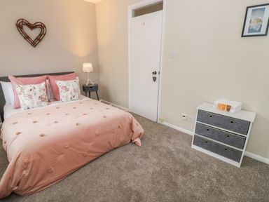 A bedroom with a bed with pillows and a pink blanket a small round bedside table with a lamp a white door a chest of drawers with toiletries and towels and a framed picture on the wall at Valentines Cottage in Stratford-Upon-Avon