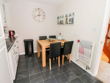 A dining area with a wooden table and four black chairs near white cabinets and a wall clock at Valentines Cottage in Stratford-Upon-Avon