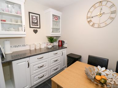 A kitchen with white cabinets a black countertop a kettle a wall clock and a dining table with a decorative fruit bowl at Valentines Cottage in Stratford-Upon-Avon