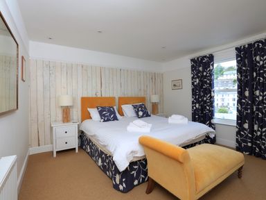 A bedroom with a double bed two bedside tables lamps a cushioned bench and a window with patterned curtains at Armorel House in Dartmouth