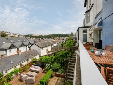 A balcony with wooden table and chairs overlooking a garden with outdoor furniture and rooftops at Armorel House in Dartmouth