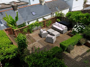 A wooden outdoor patio with cushioned seating and surrounded by various green plants and bushes at Armorel House in Dartmouth