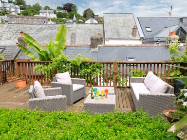 An outdoor deck with wicker furniture including two armchairs and a sofa and a table with colorful glasses and a bottle surrounded by plants at Armorel House Dartmouth