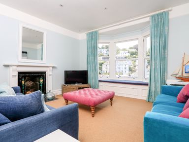 A living room with a pink ottoman, blue sofas, a fireplace, TV, and bay windows with teal curtains at Armorel House in Dartmouth