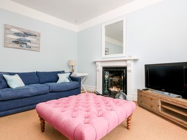 A living room with a blue sofa, pink tufted ottoman, fireplace with a mirror, wooden TV stand, and wall art at Armorel House Dartmouth